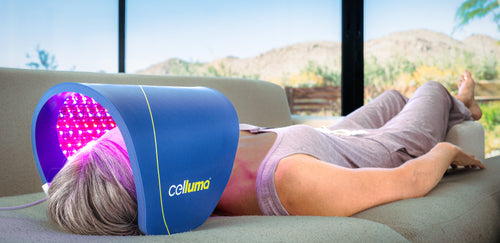 Woman lying on a couch at home under a Celluma LED light therapy device receiving a treatment for the reduction of fine lines and wrinkles. A pink glow emanates from the device that emits red, blue, and near-infrared wavelengths of light energy.